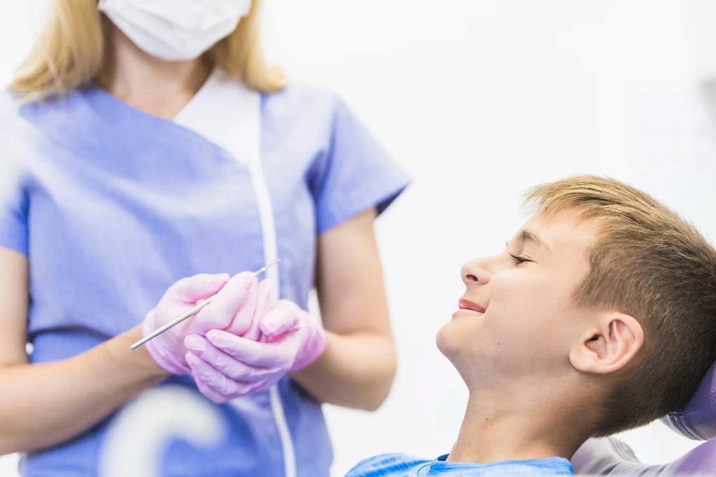 A smiling young boy sitting comfortably in a dental chair during a checkup with a female dentist wearing blue scrubs and pink gloves in San Jose.